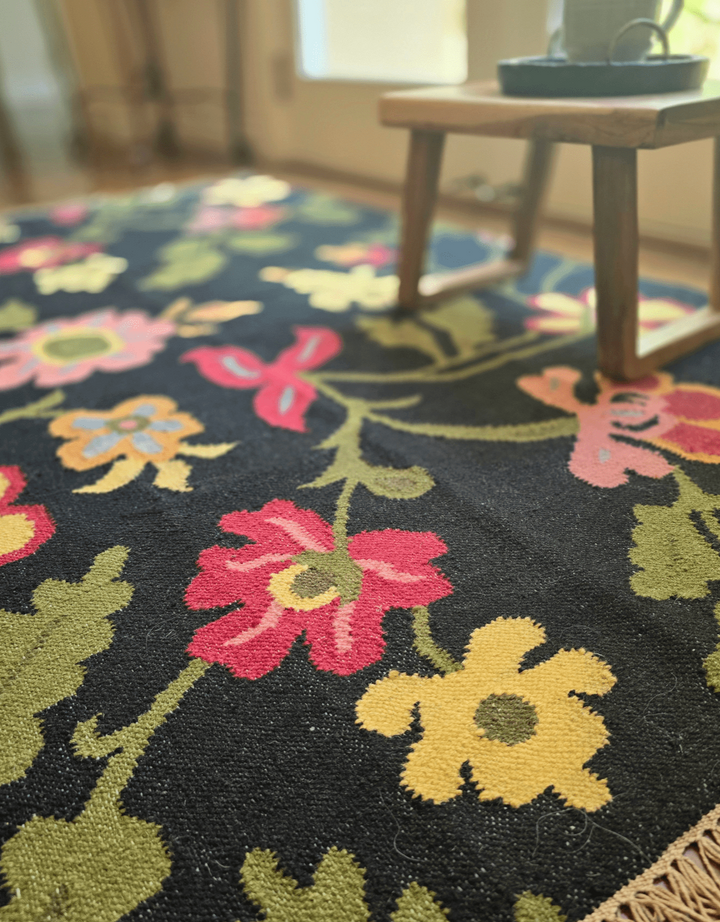 Floral patterned rug with colorful flowers on a dark background, placed in a room with a wooden chair.