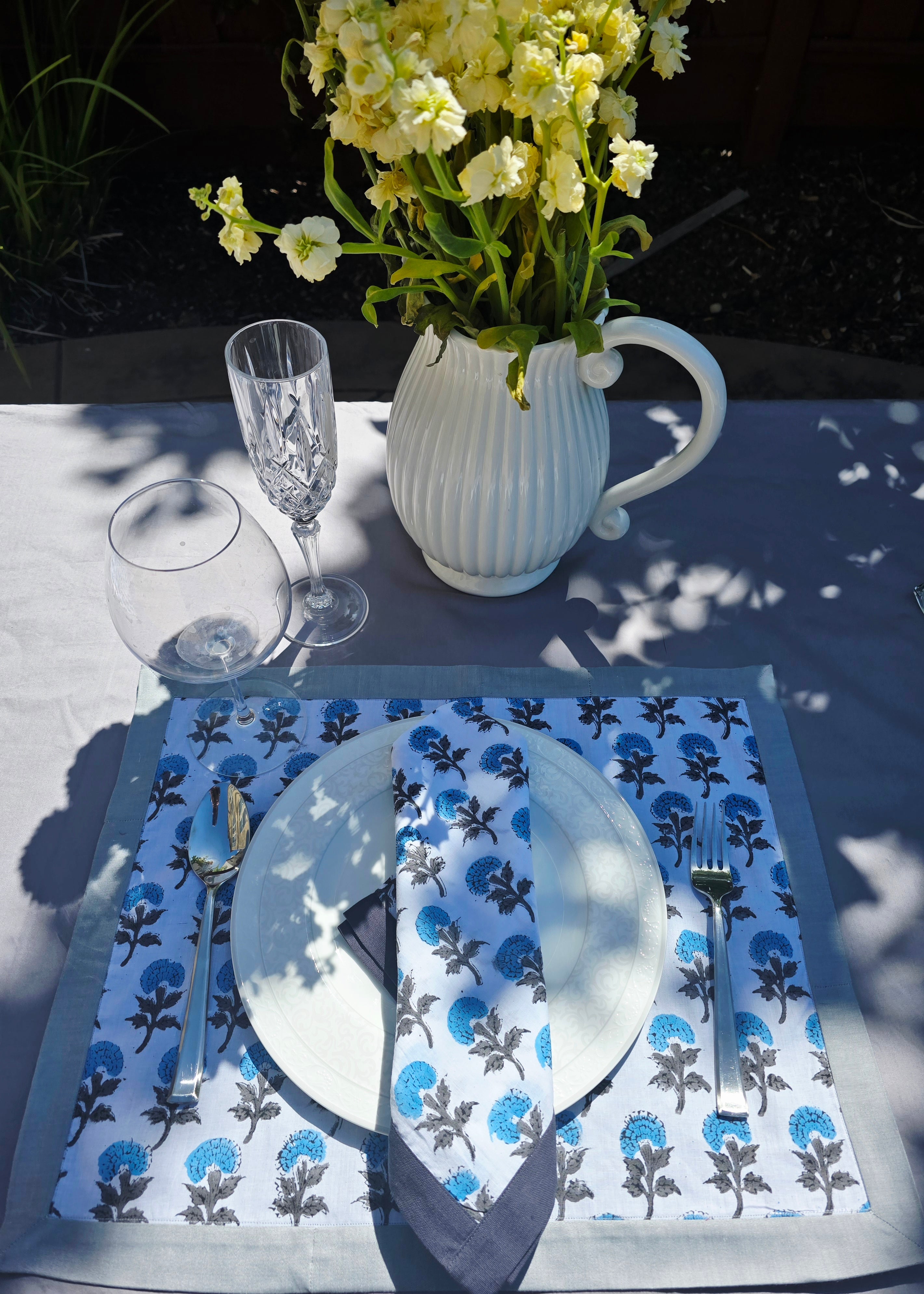 Set table with floral placemat, glassware, and vase of flowers on a sunny outdoor surface.